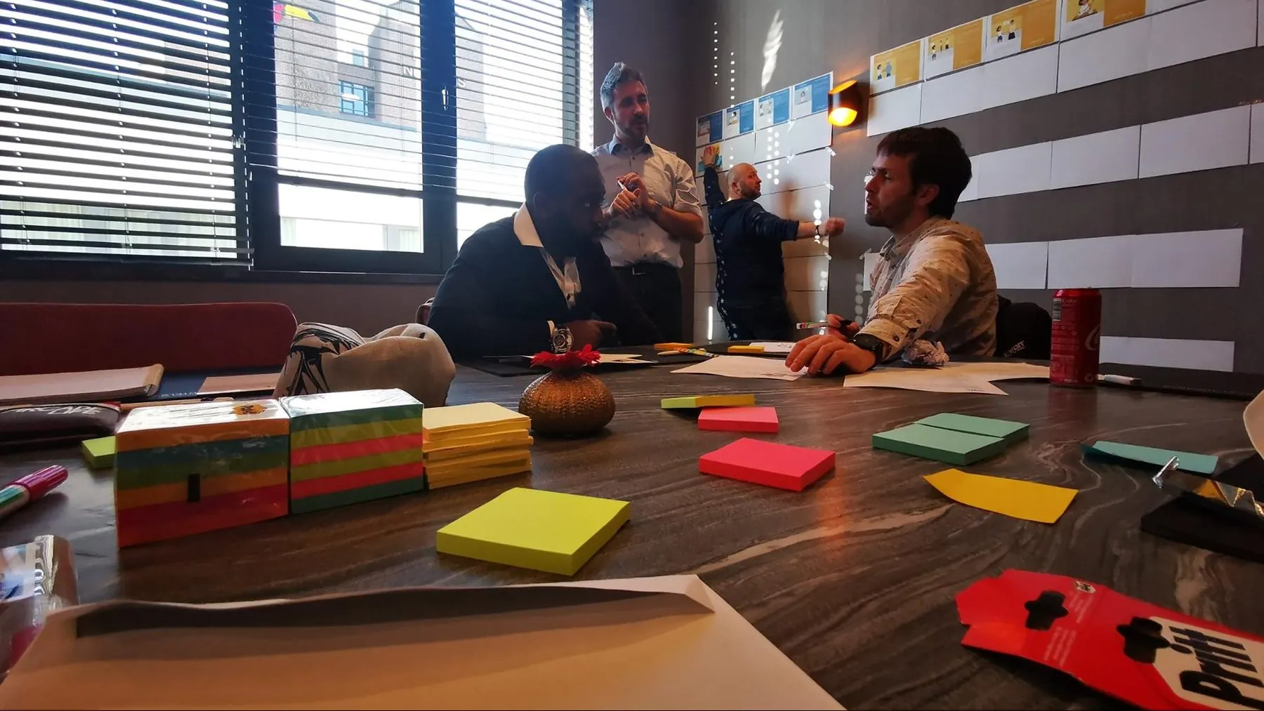 Photo of an in-person workshop: participants around a table covered in sticky notes, with personas displayed on the wall.